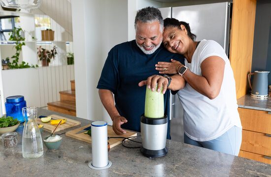 Senior couple smiling and preparing green smoothie at home kitchen island using blender and avocado