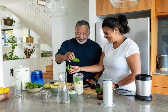 Senior African American couple preparing blended drink at kitchen island using blender jar, knife