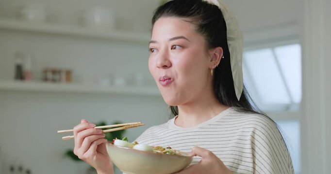 Asian woman in striped top in kitchen lifting bowl holding chopsticks tasting noodle soup chewing