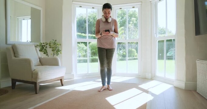 Asian woman in pink top tapping phone, sitting, placing headphones for meditation by bay window