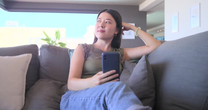 Asian adult woman checking dark blue smartphone after notification, reading messages on couch