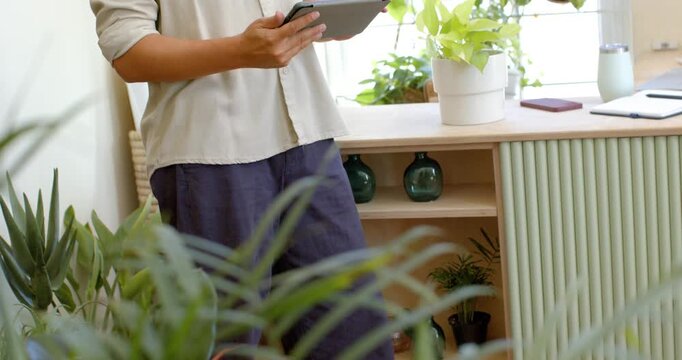 Camera revealing African nonbinary person tapping, scrolling tablet and phone for plants by window