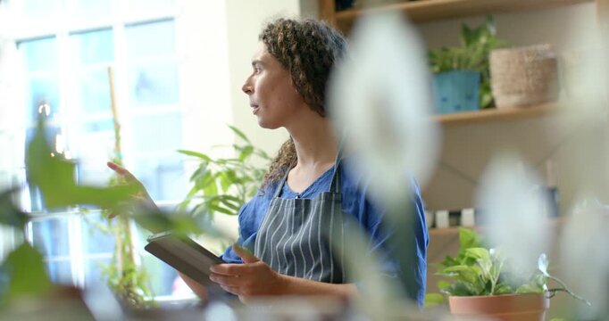 African nonbinary adult in plant shop apron glancing at window pointing tapping pad checking stock