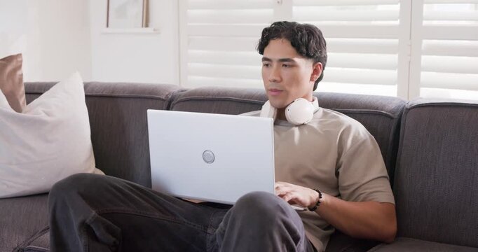 Asian man 20s reclining, sitting up, gesturing to on-screen content, typing on white laptop on sofa
