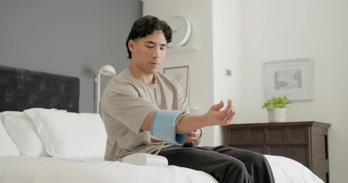 Asian man wearing beige tee reaching for blue cuff on bed, securing cuff, measuring blood pressure