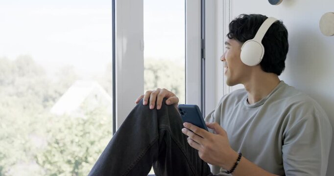 Asian man 20s checking phone wearing white headset gazing out window closing eyes winding down nook