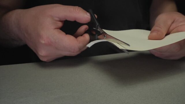 Hands using scissors to cut a piece of paper into strips on a gray surface, showcasing the precise cutting technique in a well-lit indoor setting