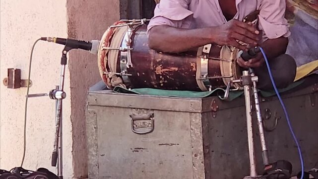 A dholak artist signals for a mic adjustment atop a metal trunk. He's preparing to amplify his instrument through loudspeakers for a public performance, his hands poised and ready to start the music.