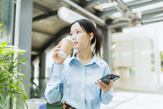 A young woman in business attire drinks coffee and uses her smartphone in a modern, sunlit indoor space.