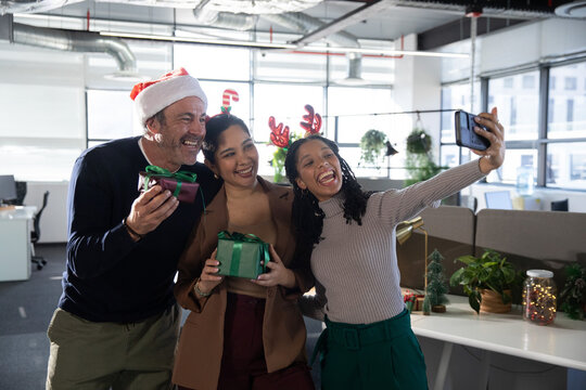 Diverse coworkers wearing Santa hat and antlers, taking selfie with smartphone and gifts in office