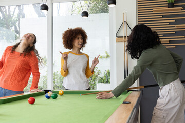Naklejka premium Three adult female friends playing pool on green table with cues and balls in recreation room