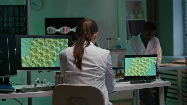 Back view of young female scientist in white coat decoding genomic sequences with microscope while sitting at table with molecular system on computer screen in laboratory