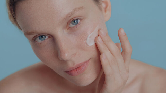 Blonde woman with bkue eyes applying facial care cream or serum on cheek and looking at camera, skincare beauty closeup portrait with a clean glowing skin