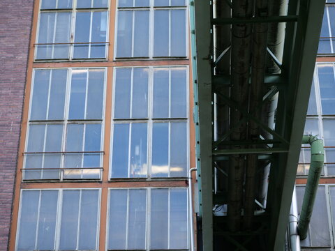 Medium shot of a weathered industrial facade featuring large multi-pane
windows with orange and green metal frames. A green industrial skybridge with
thick pipes runs vertically through 