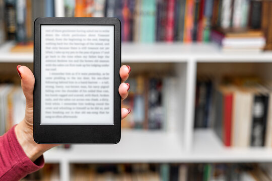a woman's hand holding an e-book. A woman reads an e-reader near a bookcase. an e-book against the background of paper books. A woman reading an e-book at home. isolate of a hand holding an e-book. 
