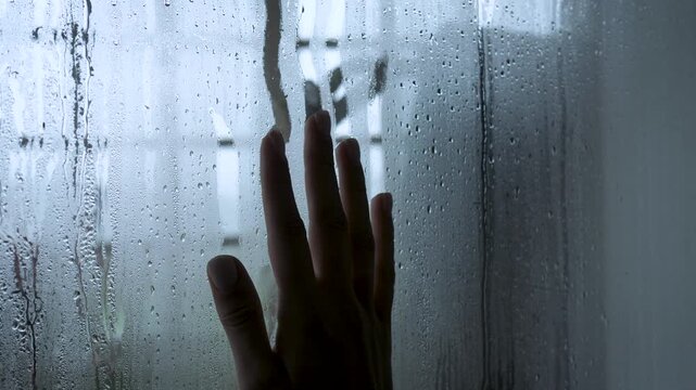 Solitary hand sliding down a steamy window covered in raindrops, creating path on condensed glass. The atmosphere is melancholic, conveying feelings of loneliness and sadness. Gesture despair of loss