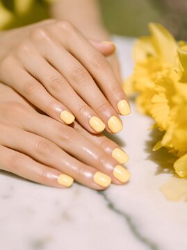 Close-up of female hands with short round yellow nails resting on a marble surface beside vibrant yellow flowers in a bright outdoor setting