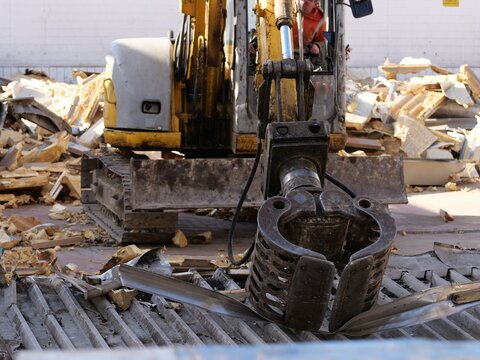 Action shot of a yellow excavator using a hydraulic sorting grapple to
grab and lift crumpled metal sheets at a demolition site. In the
background, large piles of yellow insulation foam and rubble are