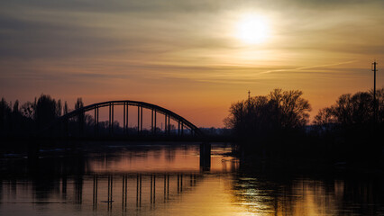 Fototapeta premium The silhouette of an old iron railway bridge with its reflection in the water in the orange rays of the setting sun.
