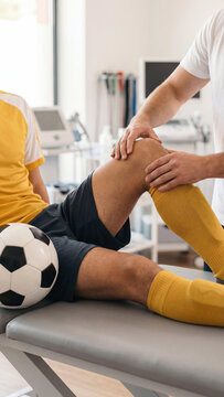 A male soccer player in a yellow jersey lying on a couch while a physiotherapist examines his injured knee in a sports clinic. Professional athlete rehabilitation concept