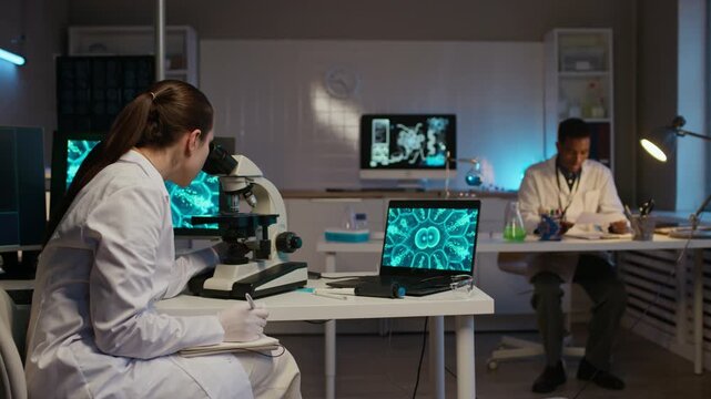 Wide stab shot of young Caucasian female microbiologist examining via microscope, taking notes and using computer software while male colleague working in background in genetics laboratory