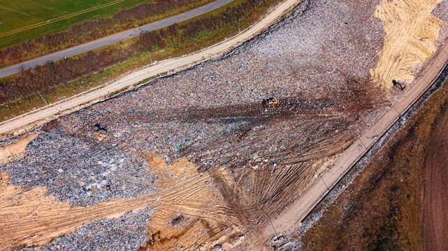 Drone view of bulldozer working on landfill pile. Aerial shot of heavy machinery compacting trash at a large waste disposal site near a road.