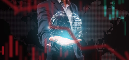 A businessman stands with a digital globe in his hand against a background of stock market trends....