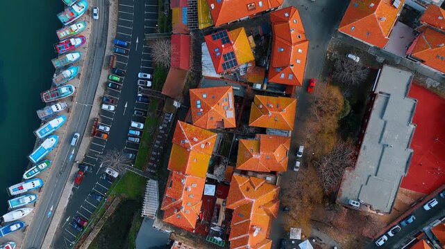 Vertical aerial view of red roofs and harbor edge. Top down shot showing a mix of residential buildings, parking and boats at the shore.
