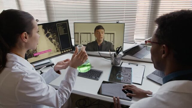 Back handheld shot of young Caucasian scientist in white coat holding flask with reagent while sitting next to Black man and video calling colleague on computer in modern clinic, discussing research