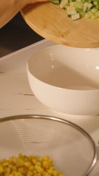 Vertical video. Woman Pouring Chopped Cucumber from Board into White Bowl, Side Angle, Fresh Salad Preparation Concept