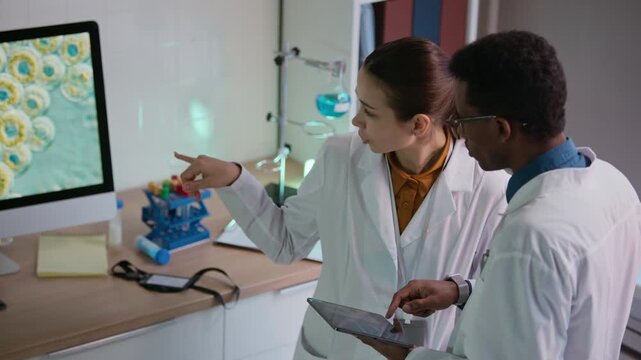 View from above of bicultural healthcare workers in white coats examining molecular system with innovative technologies using digital devices in laboratory