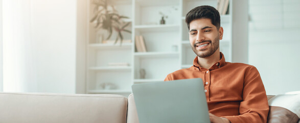 Obraz na płótnie Canvas A man sits on a couch working on a laptop. He smiles while looking at the screen. A bright room with shelves and books is in the background. Natural light fills the space.
