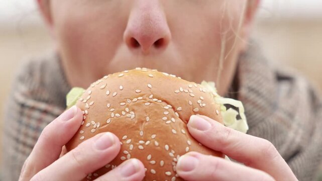 Woman eating burger outdoors, enjoying fast food during picnic in nature. Close up of hands holding burger and taking bite. Concept of casual lifestyle and comfort food.