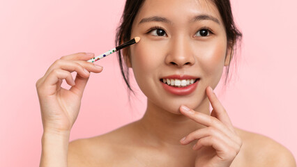 Fototapeta premium A woman holds a makeup brush near her face while smiling. She has a playful expression and is focusing on her makeup application. The background is a soft pink color.