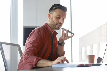 A man is sitting at a desk in an office. He is using a voice recorder while focusing on notes in a notebook. A laptop and a cup are on the table, and sunlight is coming through the window.
