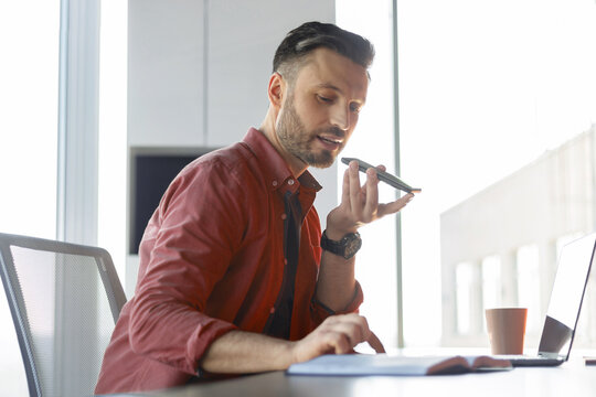 A man is sitting at a desk in an office. He is using a voice recorder while focusing on notes in a notebook. A laptop and a cup are on the table, and sunlight is coming through the window.