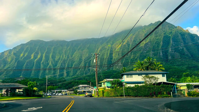 Honolulu Suburban Street with Lush Mountain Backdrop. Peaceful residential street in Honolulu with houses, palm trees, and power lines set against dramatic green mountain ridges under cloudy skies.