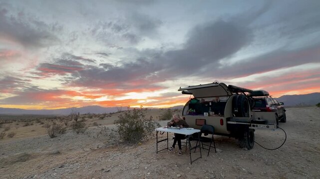 Mature woman enjoys a meal next to her teardrop camper trailer during sunset in Anza Borego Desert while overlanding.