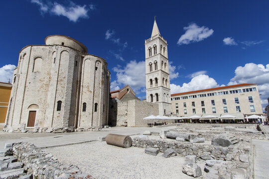 View of the Roman Forum's weathered stones contrast against the ancient cylindrical structure and the towering St. Donatus church, a timeless scene, Zadar, Zadar County, Croatia.
