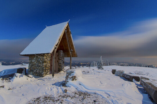 View of a snow-covered stone and wood structure atop a hill under a blue sky fading to orange at the horizon, a crisp winter scene, Schockl, Styria, Austria.
