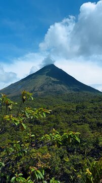 Arenal Volcano peak with clouds over lush green forest. Scenic view of the conical Arenal Volcano under a blue sky with tropical vegetation in the foreground.