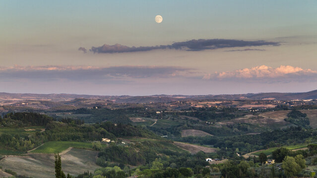 View of rolling green hills bathed in the soft glow of twilight, with a full moon hanging above, casting shadows across the landscape, San Gimignano, Tuscany, Italy.