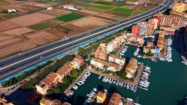 Sunlit colorful houses surrounded by waterscape and boats. Many cars move by the highways along the city. Port Saplaya, Valencia, Spain.