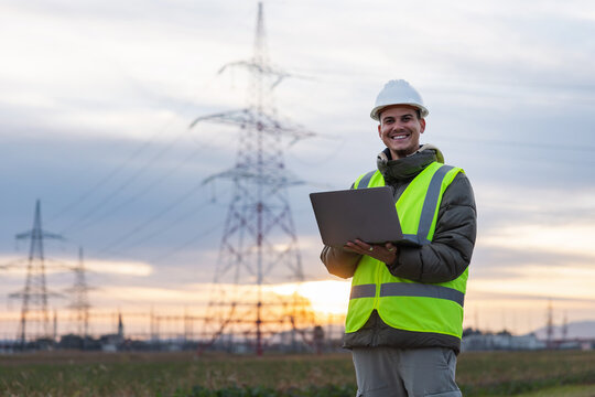 Young male electrical engineer portrait conducts inspection of high-voltage power lines using a laptop outdoors during sunset hours.