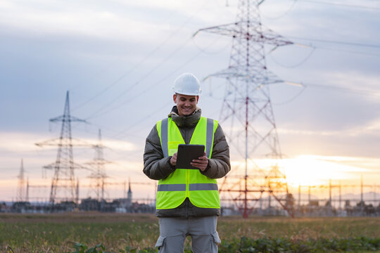 Young male electrical engineer uses a tablet to inspect high voltage lines in an outdoor area during the evening.