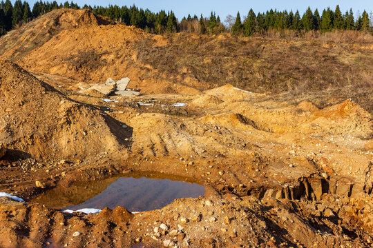 Spring thaw reveals an inactive sand quarry, forming muddy puddles among golden earthen mounds. Raw, rugged landscape under blue sky