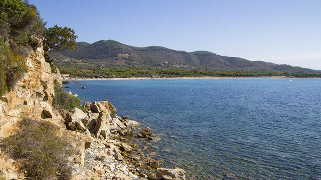 View of rugged cliffs meet the tranquil turquoise sea under a clear blue sky, as lush green hills rise in the distance, Lacona, Elba, Italy.