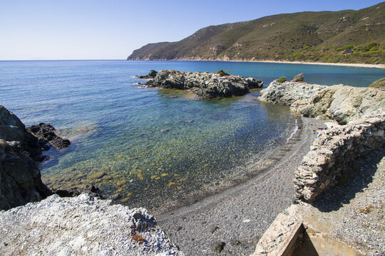 View of crystal-clear turquoise water meeting a dark pebbled shore under a vast blue sky, framed by rocky outcrops and distant green hills, Lacona, Elba, Italy.
