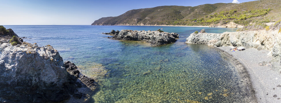 View of clear turquoise waters meeting rugged rocky shores, leading to a pebbled beach under a bright sky, in Lacona, Elba, Italy.