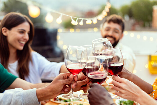 Group of diverse friends celebrating and toasting with red wine glasses during a summer party outdoor on a rooftop terrace, sharing happy moments together
